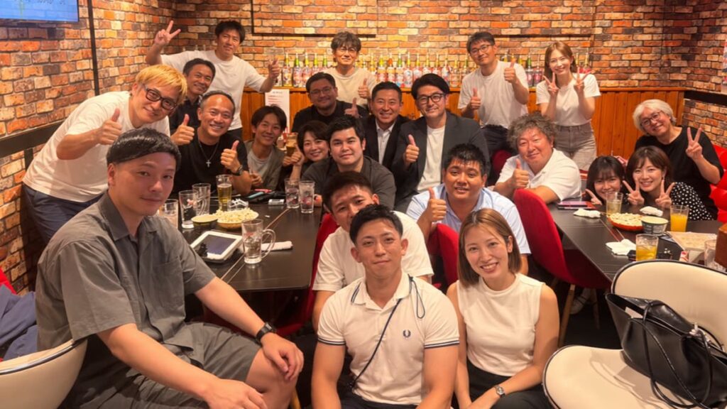 第4回愛媛県人会カラオケ交流会の集合写真。レンガ壁のバー店内で、参加者たちがテーブルを囲み、笑顔でピースサインやグッドサインをしている。